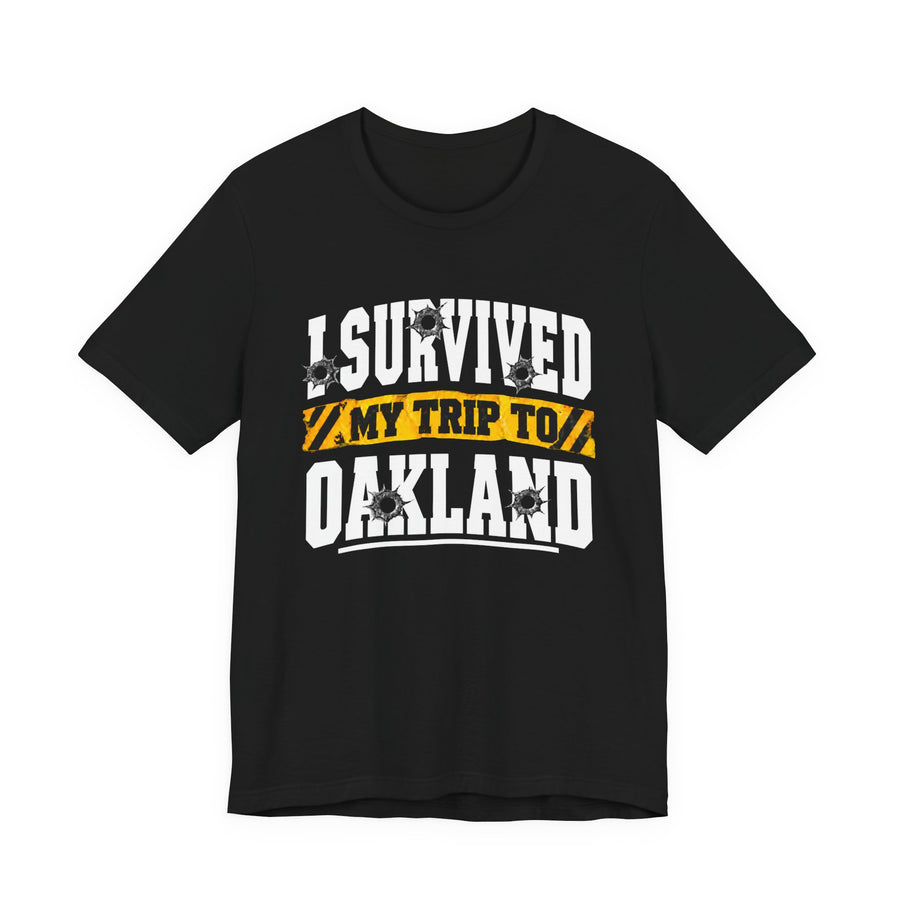 T-Shirt product photo with arched collegiate lettering reading "I SURVIVED" with bullet-hole decals, a caution-tape band reading "MY TRIP TO", and large block letters reading "OAKLAND", front view.