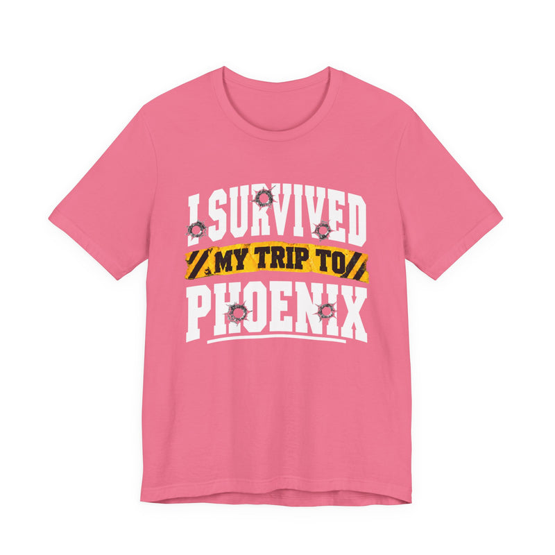 Hot pink t-shirt with white block lettering reading I SURVIVED MY TRIP TO PHOENIX, with bullet-hole accents and a yellow caution-tape banner, front flat-lay view.